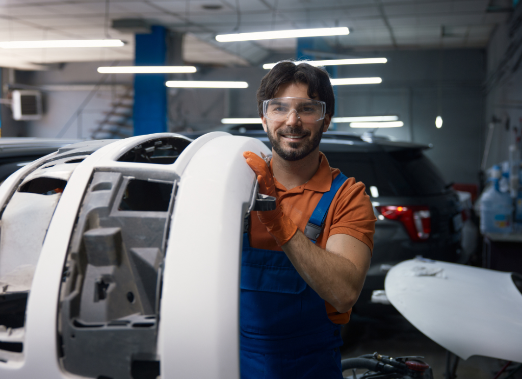 Uśmiechnięty lakiernik samochodowy w okularach ochronnych trzyma biały zderzak przygotowany do malowania w warsztacie.