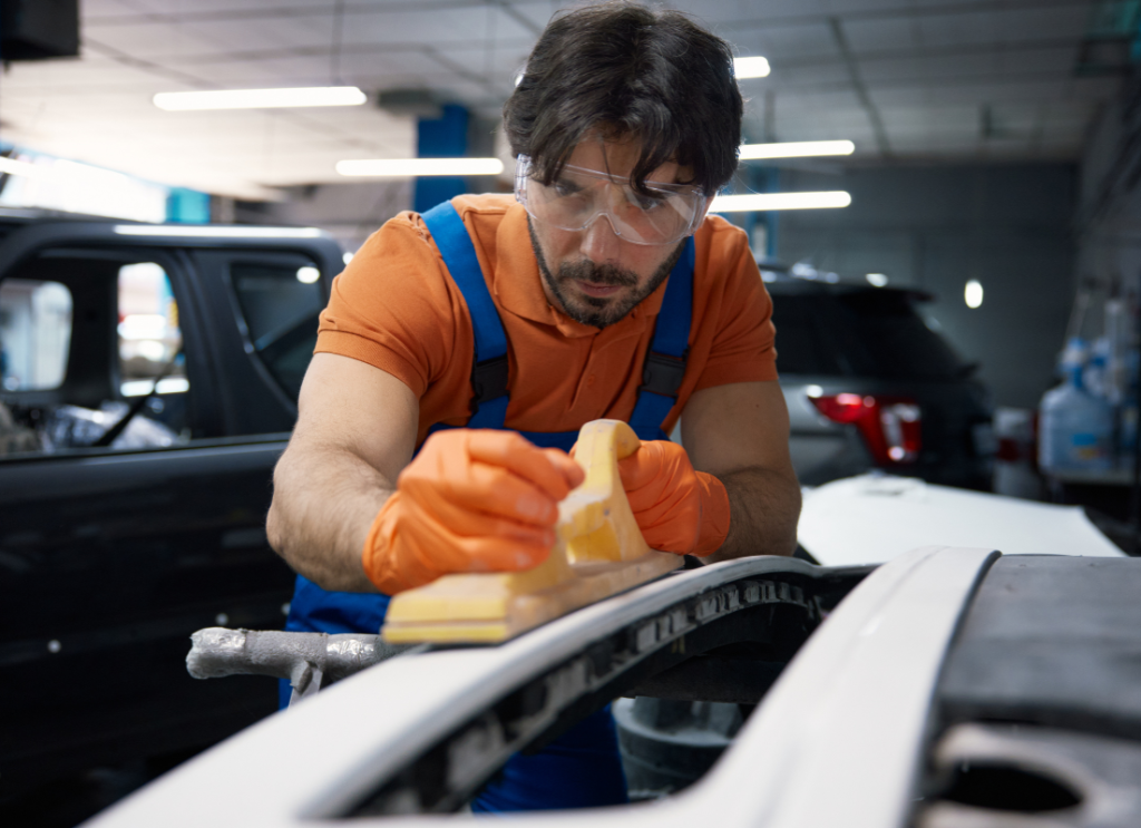 Mechanik samochodowy w okularach ochronnych ręcznie szlifuje element karoserii za pomocą klocka szlifierskiego.